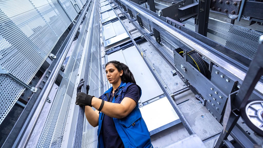 A female technician wearing a blue vest and black gloves inspects elevator cables inside a tall elevator shaft.