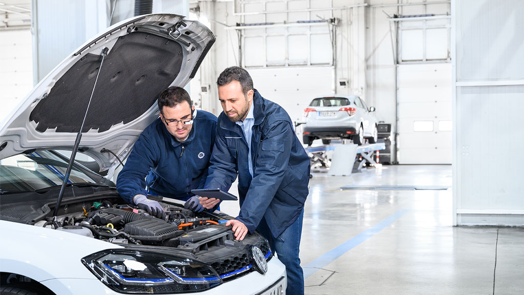 Two mechanics in blue uniforms inspect the engine of a white car with its hood open in a well-lit, spacious garage.