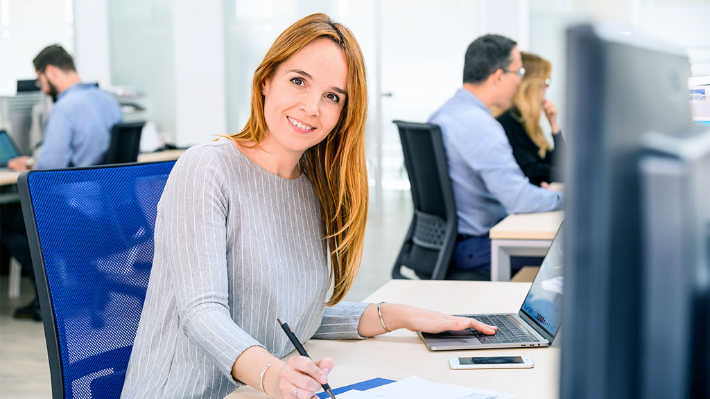 A woman in a grey sweater is sitting at a desk in a modern office, holding a pen and making notes, her other hand resting on a laptop keyboard.