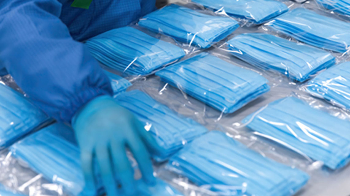 A person in blue protective clothing and gloves handles stacks of individually packaged blue surgical masks in a manufacturing facility.