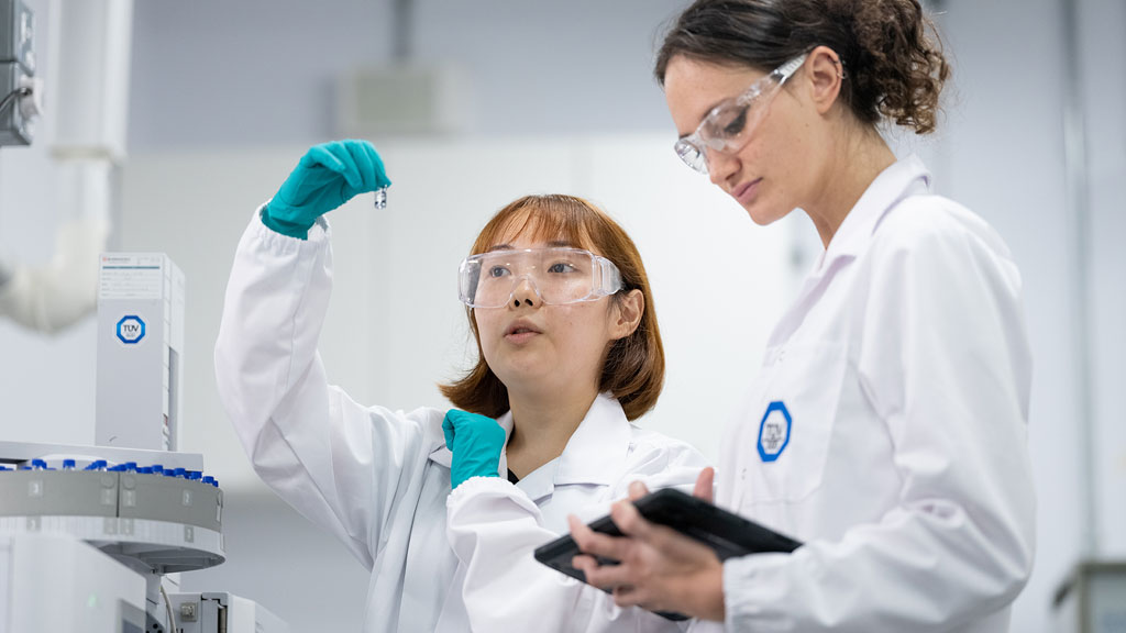 wo individuals in white lab coats and gloves work in a laboratory, with one examining a small vial or test tube and the other holding a tablet.