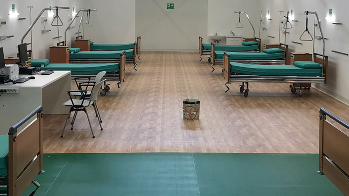 A hospital ward with multiple beds arranged in two rows, each with green bedding and medical equipment.