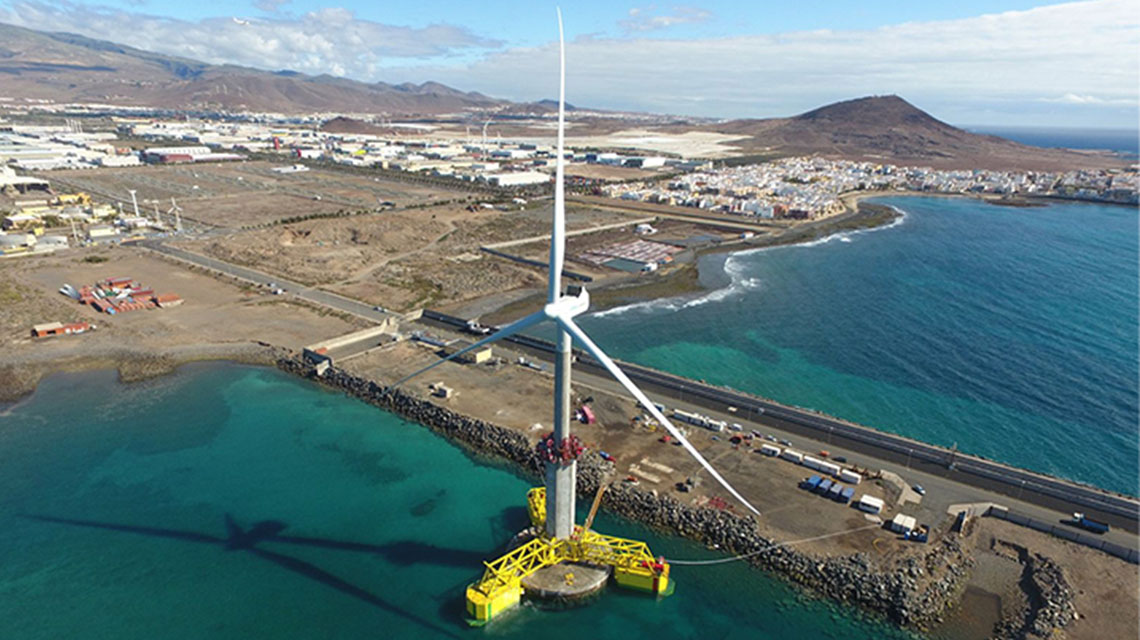 A large wind turbine situated in the water near a coastal area, mounted on a floating platform and anchored to the seabed, with an industrial zone and nearby town in the background.