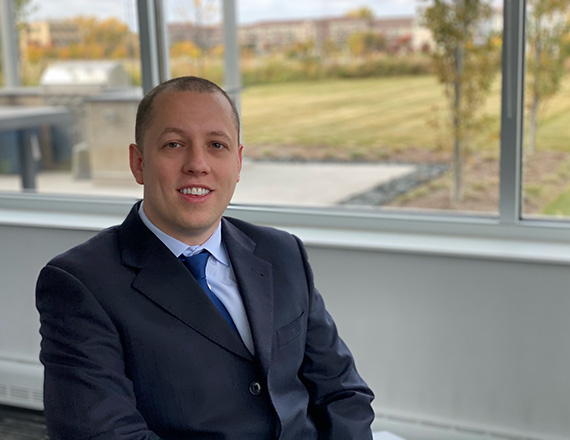 A person in a dark suit is sitting in front of a window with greenery outside.