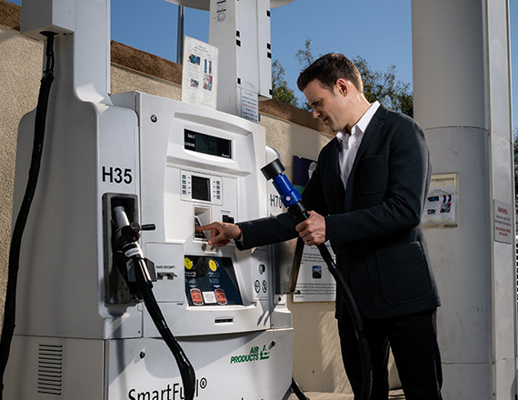A man in a suit refueling a vehicle at a hydrogen fueling station, holding the nozzle while operating the payment system.