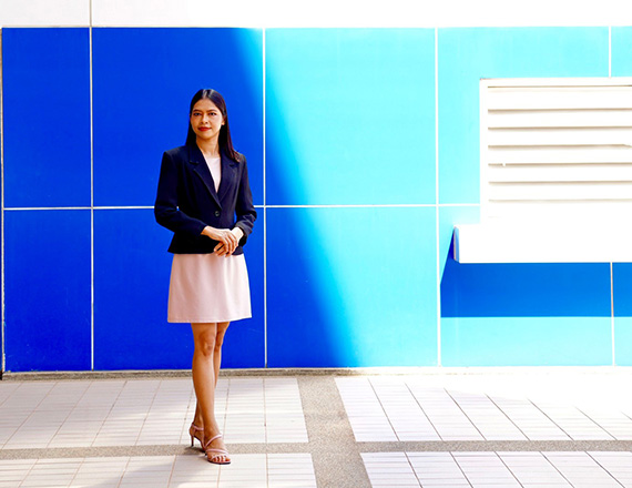 A person wearing a dark blazer and light-colored dress stands on a tiled floor in front of a vibrant blue wall.