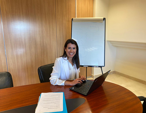 A woman sits in a small, brown conference room with a laptop in front of her and a whiteboard in the background.