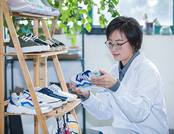 A person in a white lab coat examines a sneaker while seated next to a wooden shoe rack filled with various sneakers.