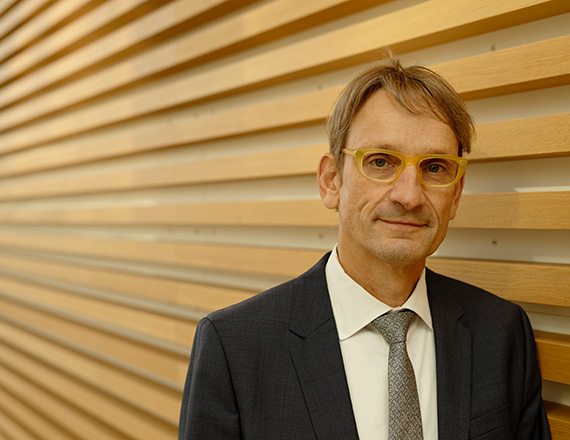 A person wearing glasses and a dark grey suit stands in front of a wall with horizontal wooden slats.
