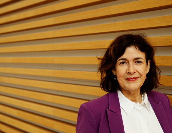 A person wearing a purple blazer over a white shirt stands in front of a wall with horizontal wooden slats.