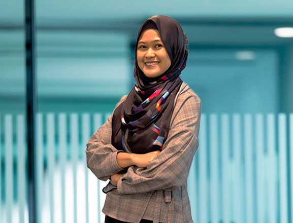 A person wearing a headscarf stands in an office corridor with glass walls.