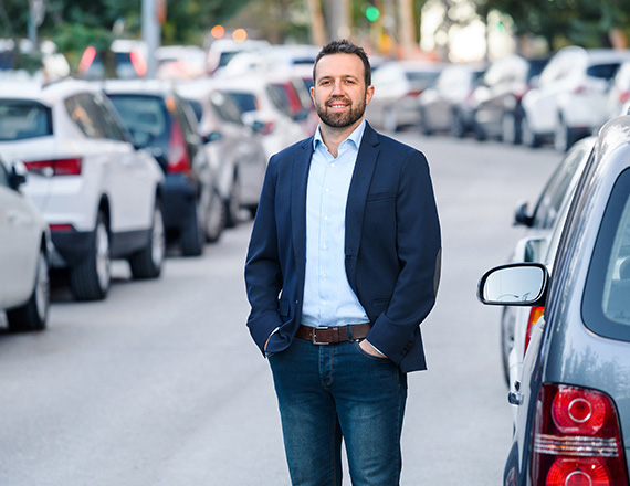 A person in a dark blazer, light blue shirt, and jeans stands on a street with parked cars on both sides.