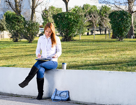 A person sitting on a low white wall in a park-like setting, writing on a notebook with a coffee cup and bag nearby.