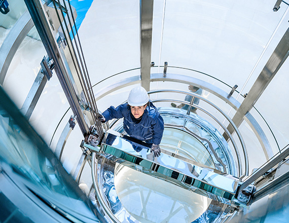 A woman in blue overalls with a white helmet on her head stands in a glass lift shaft.