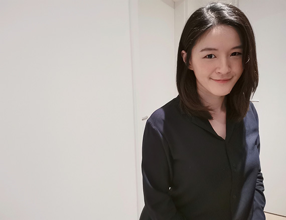 A person with shoulder-length dark hair stands against a plain white background, wearing a dark long-sleeved shirt.