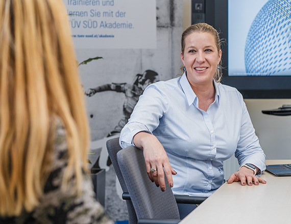 A woman in a light blue shirt is sitting at a table in an office, talking to another person.