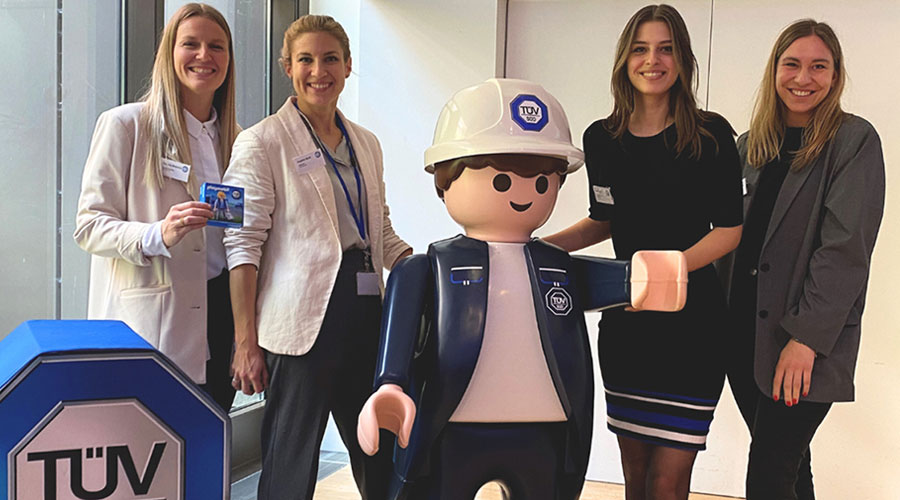 A group of young women stand smiling next to a large Playmobil figure of a TÜV SÜD inspector.
