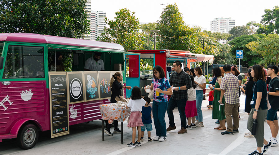 People stand in line in front of a food truck, with large green trees in the background.